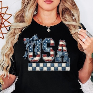 May include: Black t-shirt featuring the word "USA" in a distressed, patriotic design. The letters are styled with the American flag's colors and a denim texture, with a blue bow and a checkered border at the bottom. A casual, everyday tee.