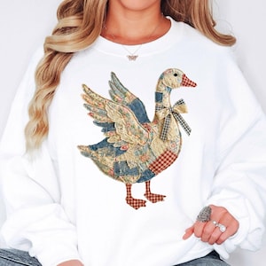 May include: White sweatshirt featuring a patchwork goose design. The goose has wings spread, a bow tie, and is made of various patterned fabrics in blue, red, and beige. The sweatshirt is a casual, comfortable style.