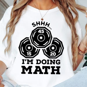 May include: White t-shirt with a black graphic featuring three weight plates with the numbers 45, 15, and 20, and the text "SHHH... I'M DOING MATH". The shirt is a casual style.