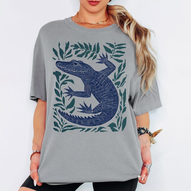 May include: A gray t-shirt featuring a navy blue alligator illustration surrounded by green leaves. The alligator is detailed with textured lines, and the leaves create a rectangular border around the design. The shirt has short sleeves and a relaxed fit.