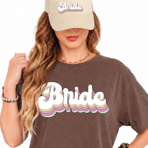May include: A dark brown t-shirt and a beige baseball cap, both featuring the word Bride in a white, retro-style font with layered pastel pink, yellow, and blue outlines. The text is printed on the center of the shirt and the front of the cap.