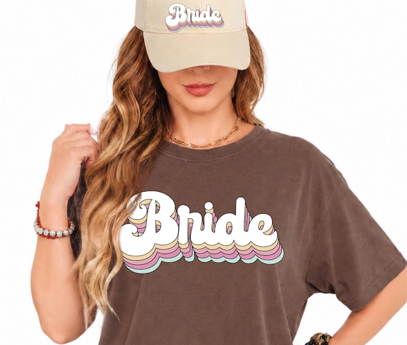 May include: A dark brown t-shirt and a beige baseball cap, both featuring the word Bride in a white, retro-style font with layered pastel pink, yellow, and blue outlines. The text is printed on the center of the shirt and the front of the cap.