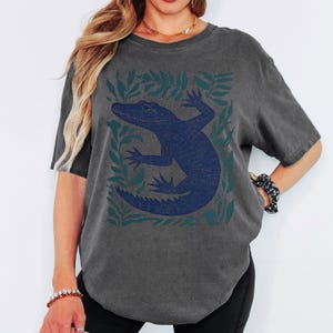 May include: Dark gray t-shirt featuring a blue alligator design surrounded by green leaves. The alligator is detailed with textured lines. The shirt has short sleeves and a relaxed fit, suitable for casual wear.