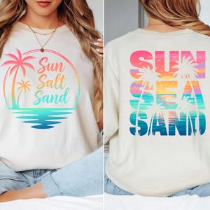 K&ouml;nnte beinhalten: Cremefarbenes T-Shirt mit farbenfroher Grafik. Auf der Vorderseite sind eine Palme, die Worte "Sun Salt Sand" in einem Regenbogenbogen und eine t&uuml;rkisfarbene Wasserlinie abgebildet. Das Design auf der R&uuml;ckseite wiederholt das Thema mit den Worten "Sun Sea Sand".