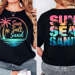 K&ouml;nnte beinhalten: Schwarzes T-Shirt mit einem farbenfrohen Aufdruck mit Palmen, den Worten "Sun Salt Sand" und einer Strandszene. Auf der R&uuml;ckseite des Shirts steht "Sun Sea Sand" in &auml;hnlichem Design.
