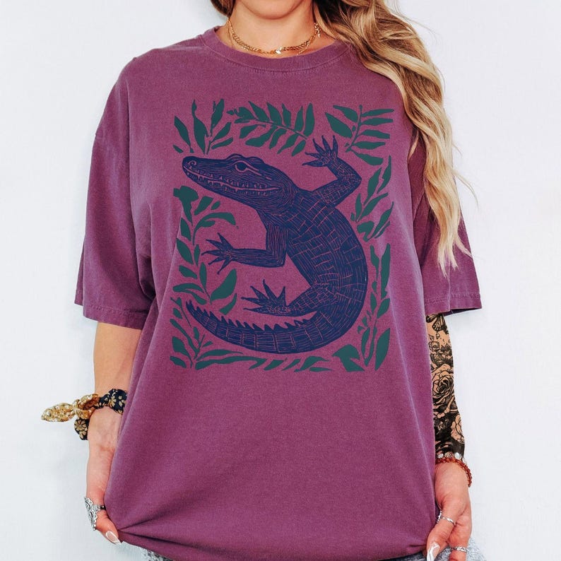 May include: A maroon t-shirt featuring a dark blue alligator illustration surrounded by green leaves. The alligator is detailed with scales and claws. The shirt has short sleeves and a relaxed fit.