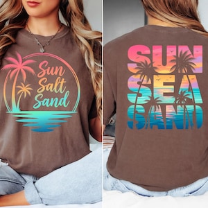 K&ouml;nnte beinhalten: Braunes T-Shirt mit einem Strandmotiv. Auf der Vorderseite befinden sich eine Palme und die Worte "Sun Salt Sand" in einem Kreis. Auf der R&uuml;ckseite steht "Sun Sea Sand" mit Palmen und einem Sonnenuntergang.