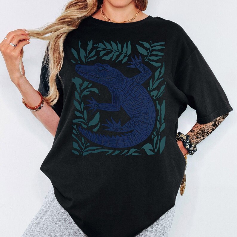 May include: Black t-shirt featuring a blue alligator design surrounded by green leaves. The alligator is detailed with textured lines, and the leaves form a square frame around the reptile. The shirt has short sleeves and a relaxed fit.