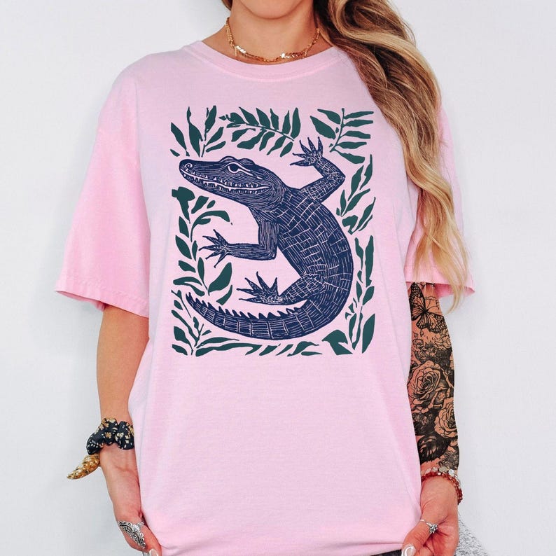 May include: A light pink t-shirt featuring a dark blue alligator illustration surrounded by green leaves. The alligator is detailed with scales and claws. The shirt has short sleeves and a relaxed fit.