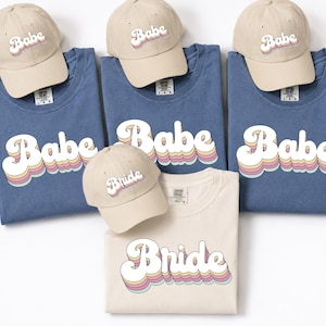 May include: Three blue t-shirts and one cream t-shirt, each featuring the word Babe or Bride in a retro-style font with layered pastel colors. Four matching cream baseball caps are included, each embroidered with the same Babe or Bride text.