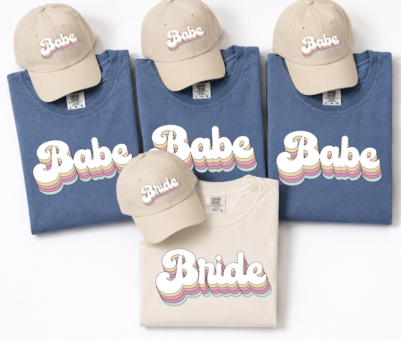 May include: Three blue t-shirts and one cream t-shirt, each featuring the word Babe or Bride in a retro-style font with layered pastel colors. Four matching cream baseball caps are included, each embroidered with the same Babe or Bride text.
