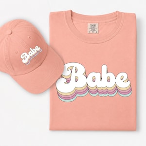 May include: A peach-colored baseball cap and a matching peach-colored t-shirt. Both items feature the word "Babe" printed in a white, retro-style font with layered, multi-colored pastel outlines in yellow, pink, and light blue.