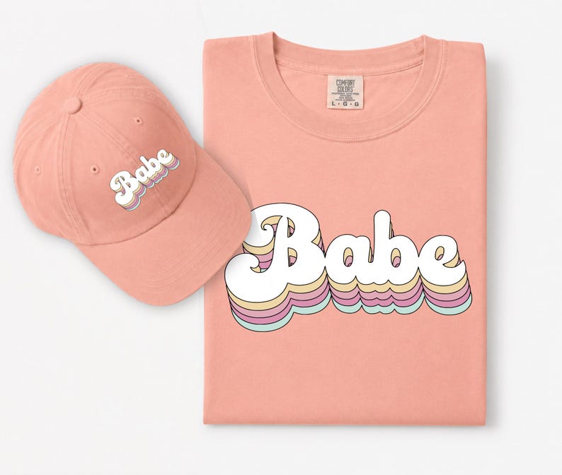 May include: A peach-colored baseball cap and a matching peach-colored t-shirt. Both items feature the word "Babe" printed in a white, retro-style font with layered, multi-colored pastel outlines in yellow, pink, and light blue.