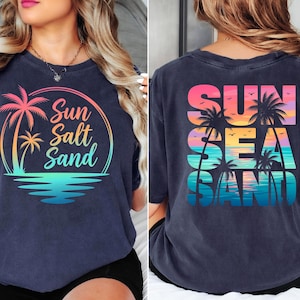 K&ouml;nnte beinhalten: Marineblaues T-Shirt mit einem farbenfrohen Grafikdesign von Palmen, den Worten "Sun Salt Sand" und einer Strandszene. Auf der R&uuml;ckseite des Shirts stehen die Worte "Sun Sea Sand" mit Palmen und einem Sonnenuntergang.