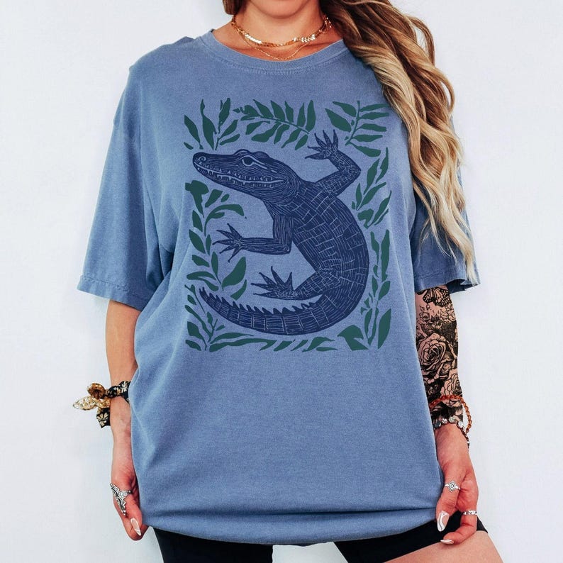 May include: A blue t-shirt featuring a dark blue alligator illustration surrounded by green leaves. The alligator is detailed with textured lines. The shirt has short sleeves and a relaxed fit, suitable for casual wear.