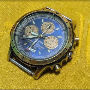 May include: A round analog wristwatch with a metallic gold-tone case and a blue dial. The watch features three sub-dials, luminous hands, and a tachymeter scale on the bezel. Text on the dial includes Field Gear, Alarm Chronograph, and Water Resist.