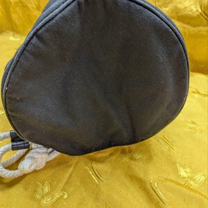May include: A black, cylindrical drawstring bag with a rounded top. The bag is made of a dark fabric and features a white rope drawstring. The bag is resting on a gold-colored surface.