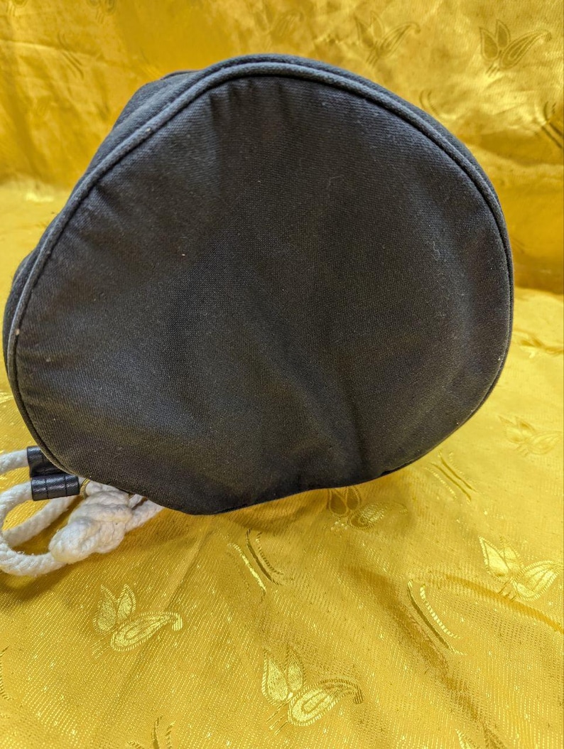 May include: A black, cylindrical drawstring bag with a rounded top. The bag is made of a dark fabric and features a white rope drawstring. The bag is resting on a gold-colored surface.