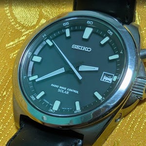 May include: A Seiko watch with a dark green face and silver casing. The watch has white hour markers and hands, with the text "SEIKO" and "RADIO WAVE CONTROL SOLAR". The watch has a black leather strap.