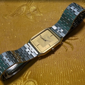 May include: A gold-toned rectangular wristwatch with a silver and green metal band. The watch face is a light gold color with black hands and markers. The band is made of interlocking metal links.