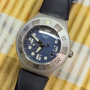 May include: A Swatch wristwatch featuring a dark blue dial with white numerals at the 3, 6, and 9 o'clock positions, luminous hands, and circular hour markers. The watch has a silver-toned bezel with black engraved numbers and a black strap.