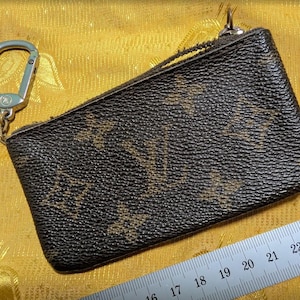 May include: A brown and tan coin purse with a zipper closure and a silver-colored carabiner clip. The purse has a textured surface with a repeating pattern. A ruler is visible in the image.