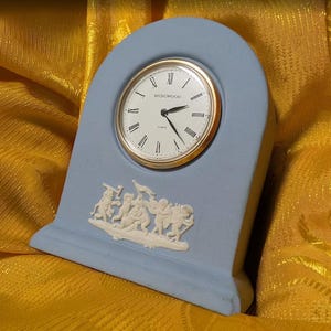 May include: A light blue arched mantel clock with a white face, gold-tone bezel, and black Roman numerals. The word Wedgwood is printed on the clock face. A white relief sculpture featuring several figures is centered on the lower front section.