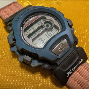 Può includere: Un orologio digitale G-Shock blu con cinturino nero e rosa. Il quadrante mostra l'ora e la data. Sul cinturino è stampata la scritta "X-treme".