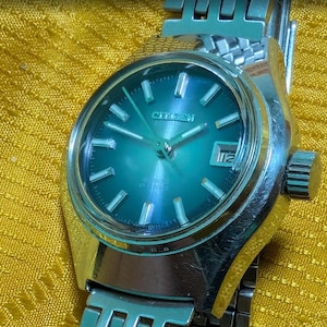 May include: A vintage Citizen watch with a teal-colored face and silver-tone metal band. The watch face features silver hour markers and hands, with a date display at the 3 o'clock position. The watch is set against a yellow background.