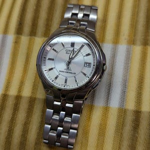 Könnte beinhalten: Eine silberfarbene, funkgesteuerte Citizen Eco-Drive Attesa Armbanduhr mit Metallgliederarmband. Die Uhr verfügt über ein rundes silbernes Zifferblatt mit silbernen Stundenmarkierungen, silbernen Zeigern, einem Datumsfenster bei drei Uhr und Text.