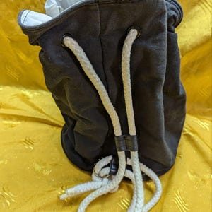 May include: A dark gray drawstring bag with a white interior and white rope straps. The bag is made of a sturdy fabric and features metal grommets for the drawstring. The rope straps are held together with a black leather strap.