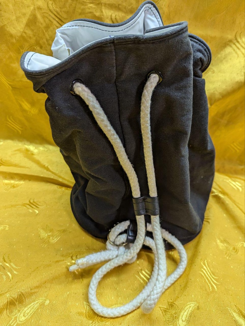 May include: A dark gray drawstring bag with a white interior and white rope straps. The bag is made of a sturdy fabric and features metal grommets for the drawstring. The rope straps are held together with a black leather strap.