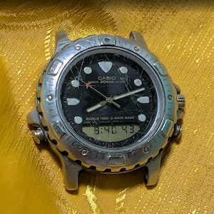 May include: A silver-tone Casio analog-digital watch with a black face, featuring the text CASIO, WATER 20BAR RESIST, and WORLD TIME & DATA BANK. The watch has a scratched crystal, luminous hour markers, and a digital display showing 8:40 43.