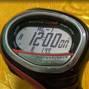 May include: A black and red SOMA LAP 300 digital watch displaying the time 12:00. The watch has a silver bezel and buttons for adjust, mode, start/stop, next, and lap/save. The watch face also displays "AL-1" and "1:49".