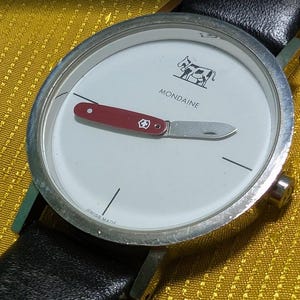May include: A wristwatch with a silver-tone round case and a black leather strap. The white dial features the text MONDAINE, a black cow graphic, and the text SWISS MADE. The hour and minute hands are shaped like a small red Swiss Army pocket knife.