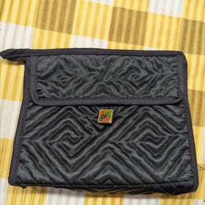 May include: Black quilted handbag with a geometric design. The bag features a flap closure with a small, colourful emblem. It has a handle on top and is set against a yellow and white striped background.