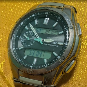 May include: A silver Casio Multi Band 6 watch with a black face. The watch displays the time digitally and with hands. The watch has a silver band and a variety of buttons and dials. The watch is on a yellow surface.