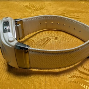 May include: A white digital wristwatch with a cream-colored canvas and leather band. The watch features a rectangular face with silver-tone metal accents. The band has multiple adjustment holes.
