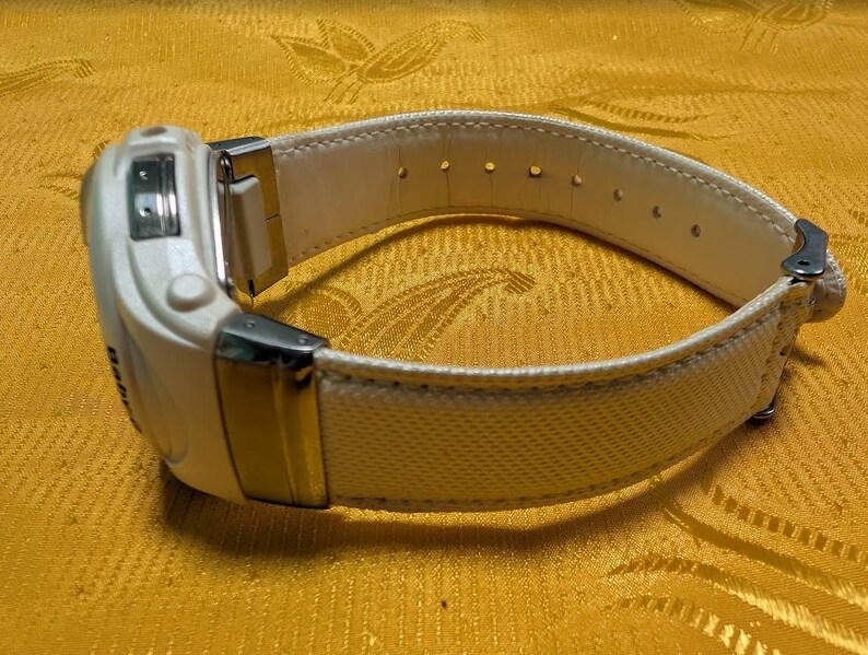 May include: A white digital wristwatch with a cream-colored canvas and leather band. The watch features a rectangular face with silver-tone metal accents. The band has multiple adjustment holes.