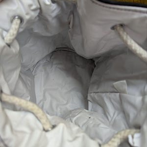 May include: A white drawstring bag with a quilted interior. The bag has a black trim and white rope drawstrings. The interior is visible, showing the quilted fabric and the drawstring closure.