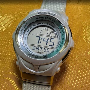 Puede incluir: Un reloj digital Baby-G Wave Ceptor blanco con bisel verde claro. La esfera muestra la hora 7:45 y la fecha SAT56. Incluye resistencia a golpes y al agua.