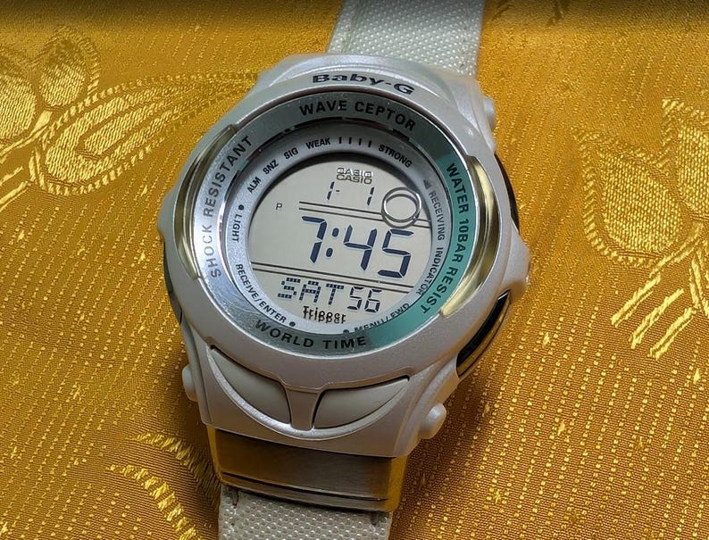May include: A white Baby-G Wave Ceptor digital watch with a light green bezel. The watch face displays the time as 7:45 and the date as SAT56. Features include shock resistance and water resistance.