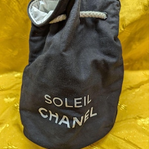 May include: Black drawstring bag with the text "SOLEIL CHANEL" in white. The bag has a white interior and a white drawstring closure. The bag is made of a dark fabric.