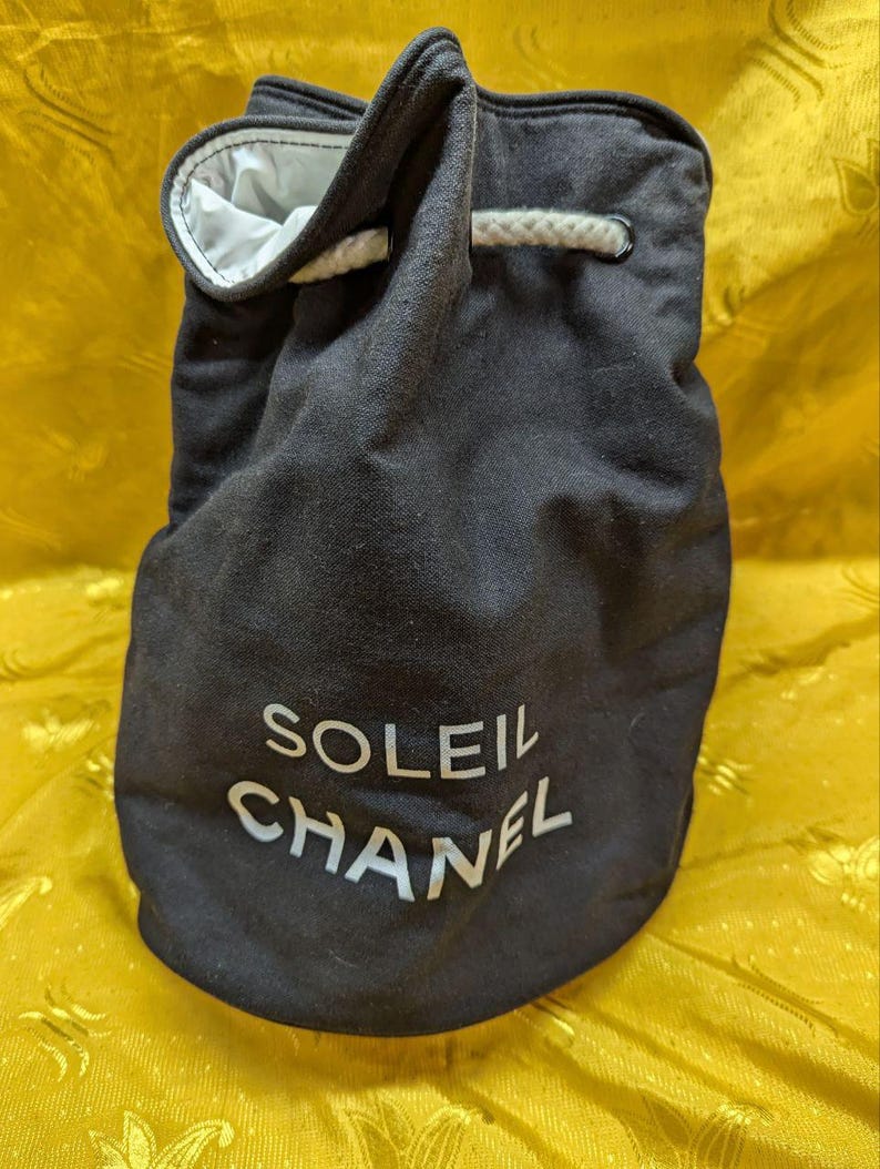 May include: Black drawstring bag with the text "SOLEIL CHANEL" in white. The bag has a white interior and a white drawstring closure. The bag is made of a dark fabric.