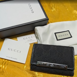 May include: A black Gucci key case with a silver-tone metal bar, presented with a white dust bag, box, and authentication cards. The key case features a patterned design.