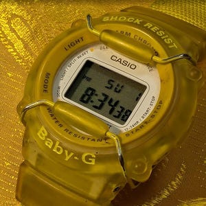 Puede incluir: Un reloj digital Baby-G amarillo con una esfera blanca y una pantalla digital negra que muestra la hora. El reloj tiene una correa amarilla y es resistente al agua. La esfera del reloj dice "CASIO" y "SHOCK RESIST".