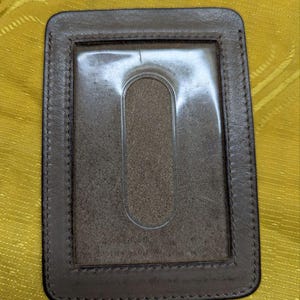 May include: A brown leather ID card holder with a clear plastic window and a stitched border. The holder has a rectangular shape with rounded corners and a vertical oval cutout.
