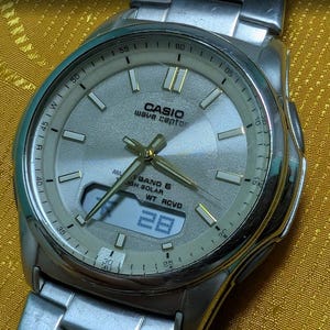 Puede incluir: Un reloj Casio Wave Ceptor plateado con esfera gris claro. El reloj tiene una pantalla digital que muestra el número 28. Las manecillas y los marcadores de hora son dorados.