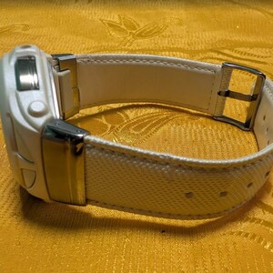 May include: A white digital watch with a textured white band and a silver buckle. The watch face has a rectangular shape with a few buttons. The band is secured with a silver buckle.