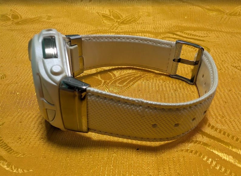 May include: A white digital watch with a textured white band and a silver buckle. The watch face has a rectangular shape with a few buttons. The band is secured with a silver buckle.